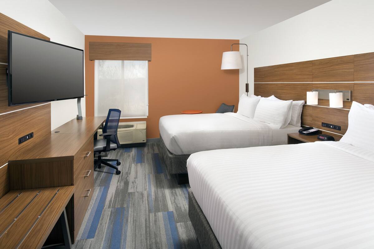 A hotel room with two beds, a desk, a TV, and a window with blinds.