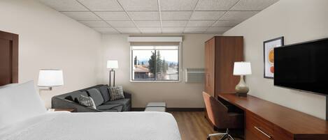 A hotel room with a bed, a desk, a chair, a television, and a window with a view of trees and mountains.