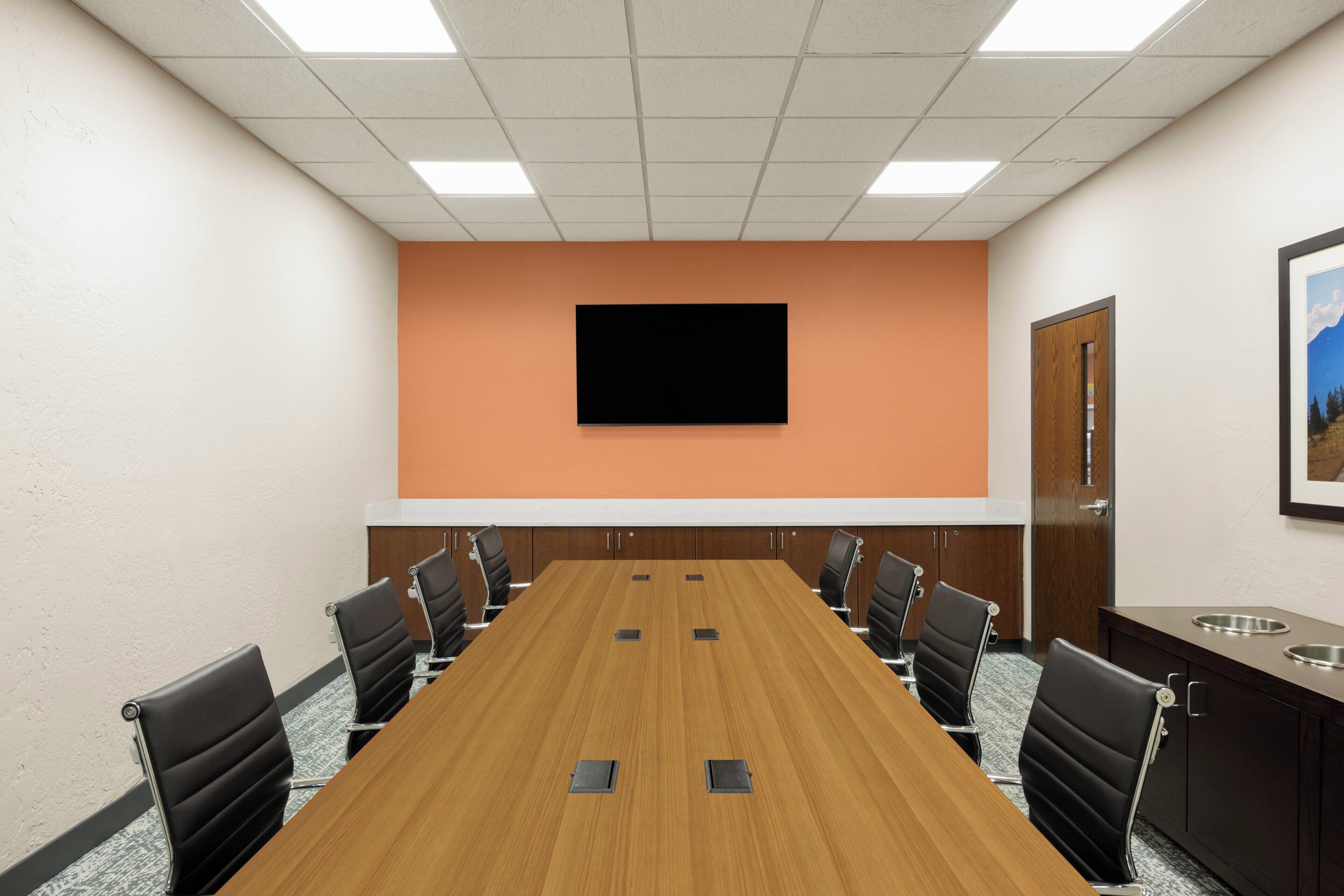 Un salón de conferencias con una larga mesa, sillas negras, un televisor montado y una puerta que da a vistas del exterior.