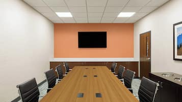 Un salón de conferencias con una larga mesa, sillas negras, un televisor montado y una puerta que da a vistas del exterior.
