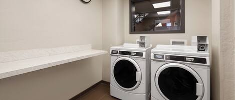 A laundry room with two front-loading washing machines, a wall-mounted clock, and a window with a view of the outside.