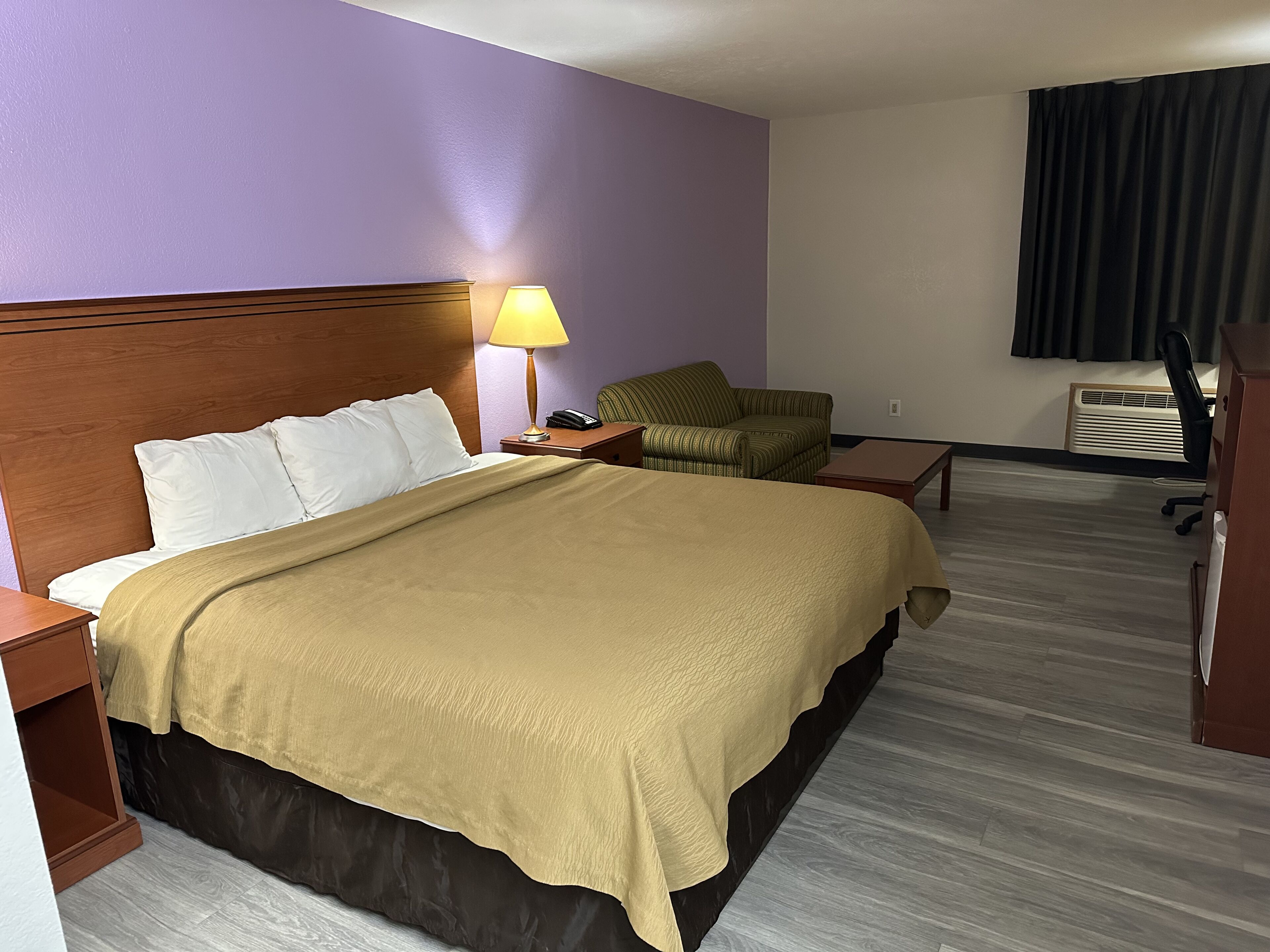 A hotel room with a large bed, a bedside lamp, a chair, a desk, and a window with curtains.