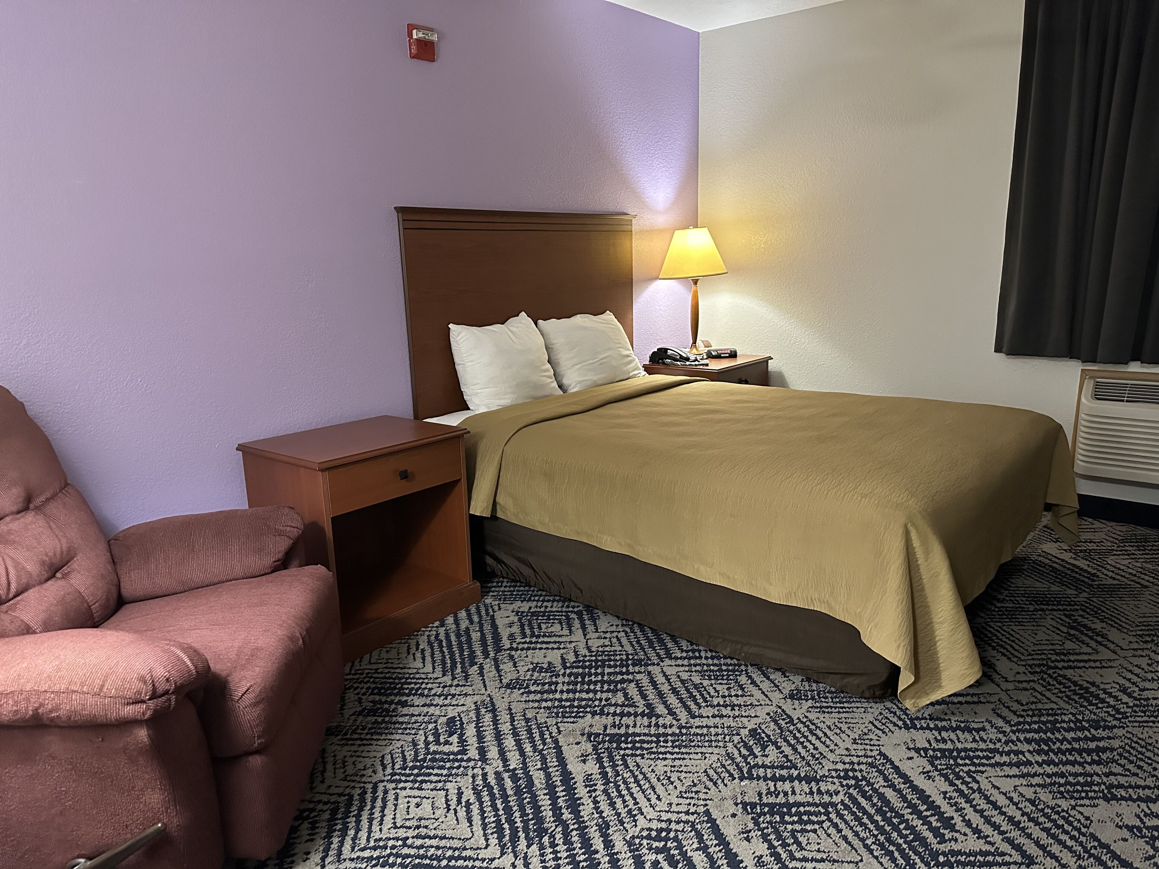 A hotel room with a bed, a nightstand, a lamp, a chair, and a patterned carpet.