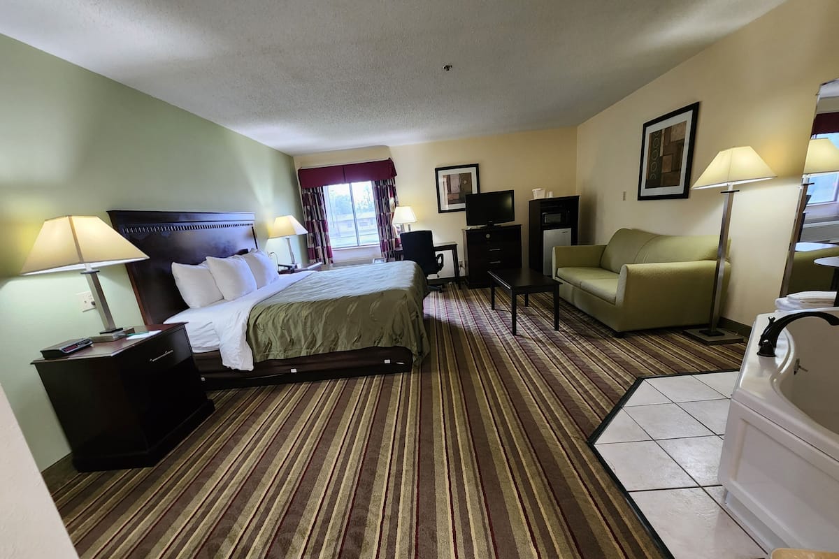 A hotel room with a bed, a desk, a chair, a TV, a small table, and a bathroom.