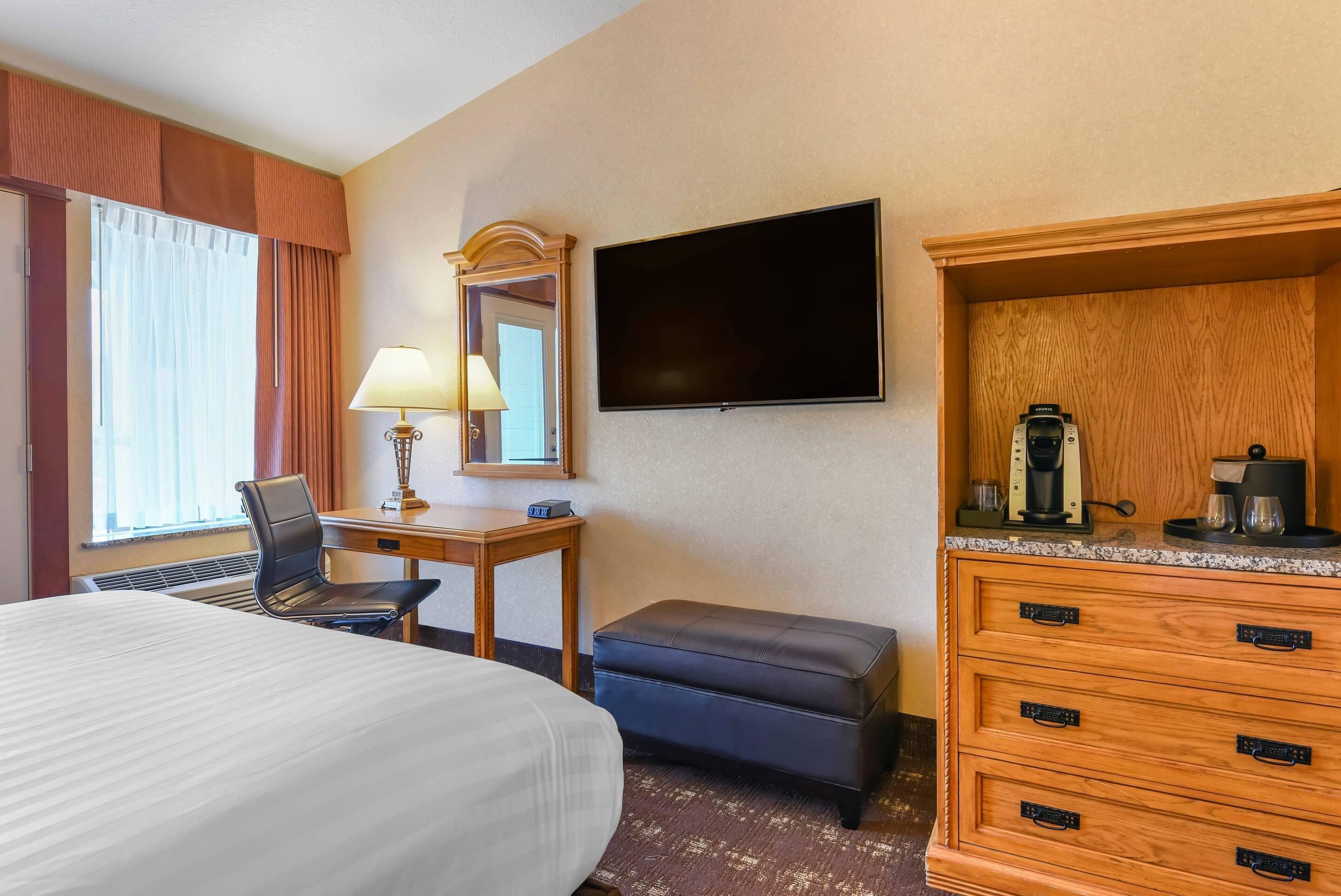 A hotel room with a large bed, a desk with a lamp, a television, a wooden dresser, and a window with curtains.