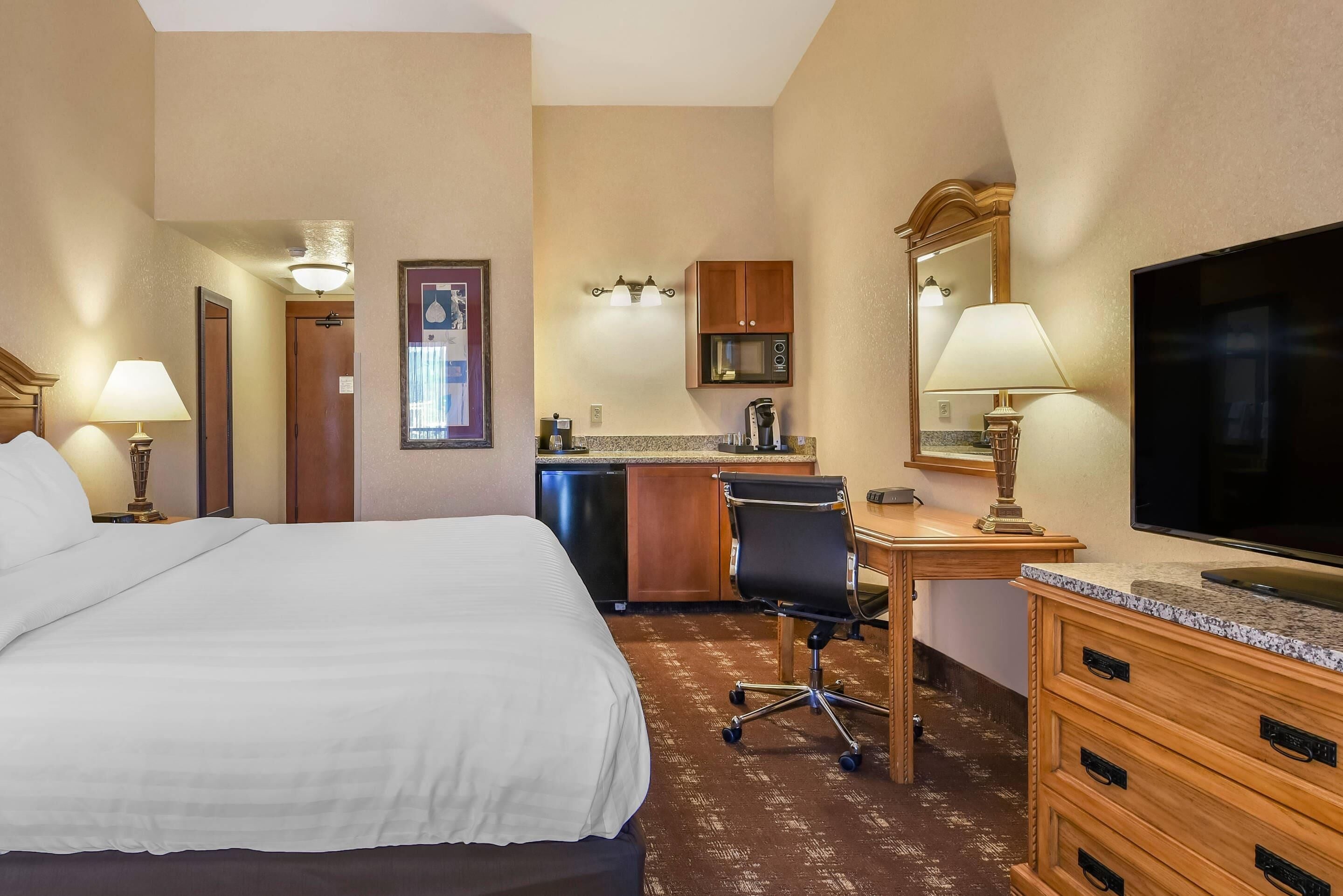 A hotel room with a bed, desk, chair, kitchenette, and a TV.