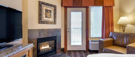 Habitación de hotel con televisión de pantalla plana, chimenea, cama, sofá de cuero, lámpara y puerta con vistas.