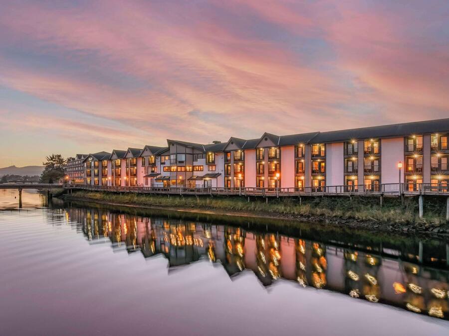 A row of modern, multi-story hotel buildings with balconies, reflecting in a calm body of water.