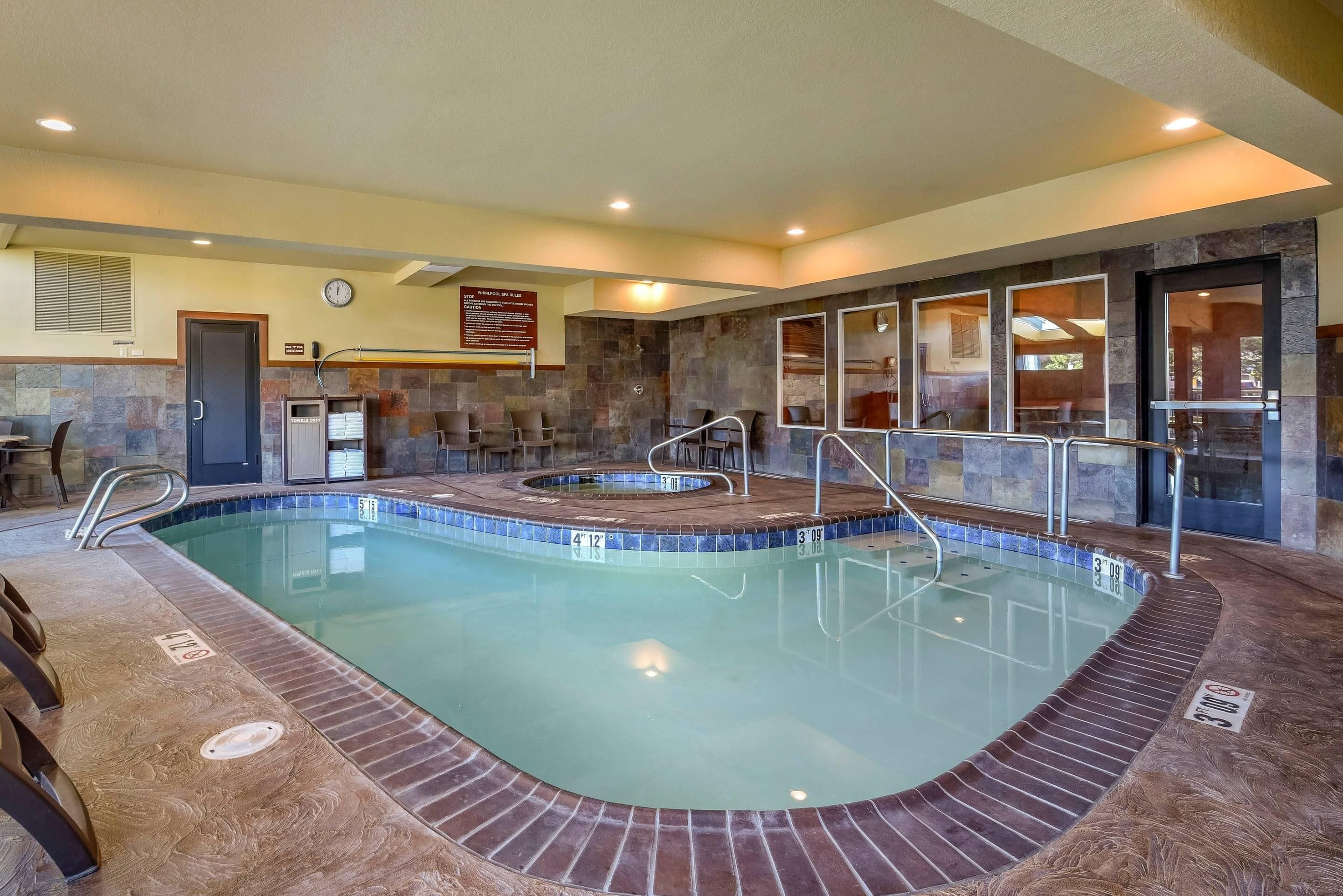 Une piscine intérieure dont le bord est revêtu de pierres, un jacuzzi et un espace détente avec des fauteuils.