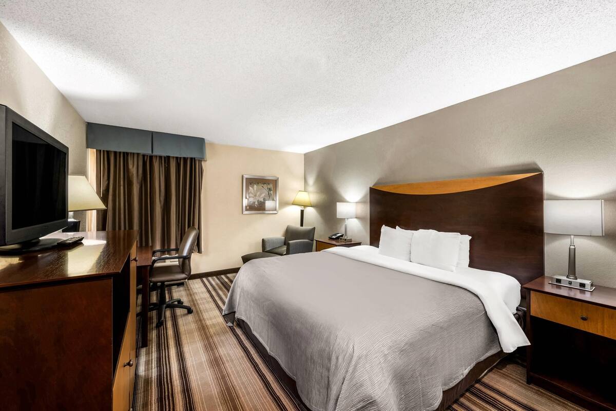 A hotel room with a large bed, a desk with a television, a chair, a desk lamp, and a nightstand with a lamp.