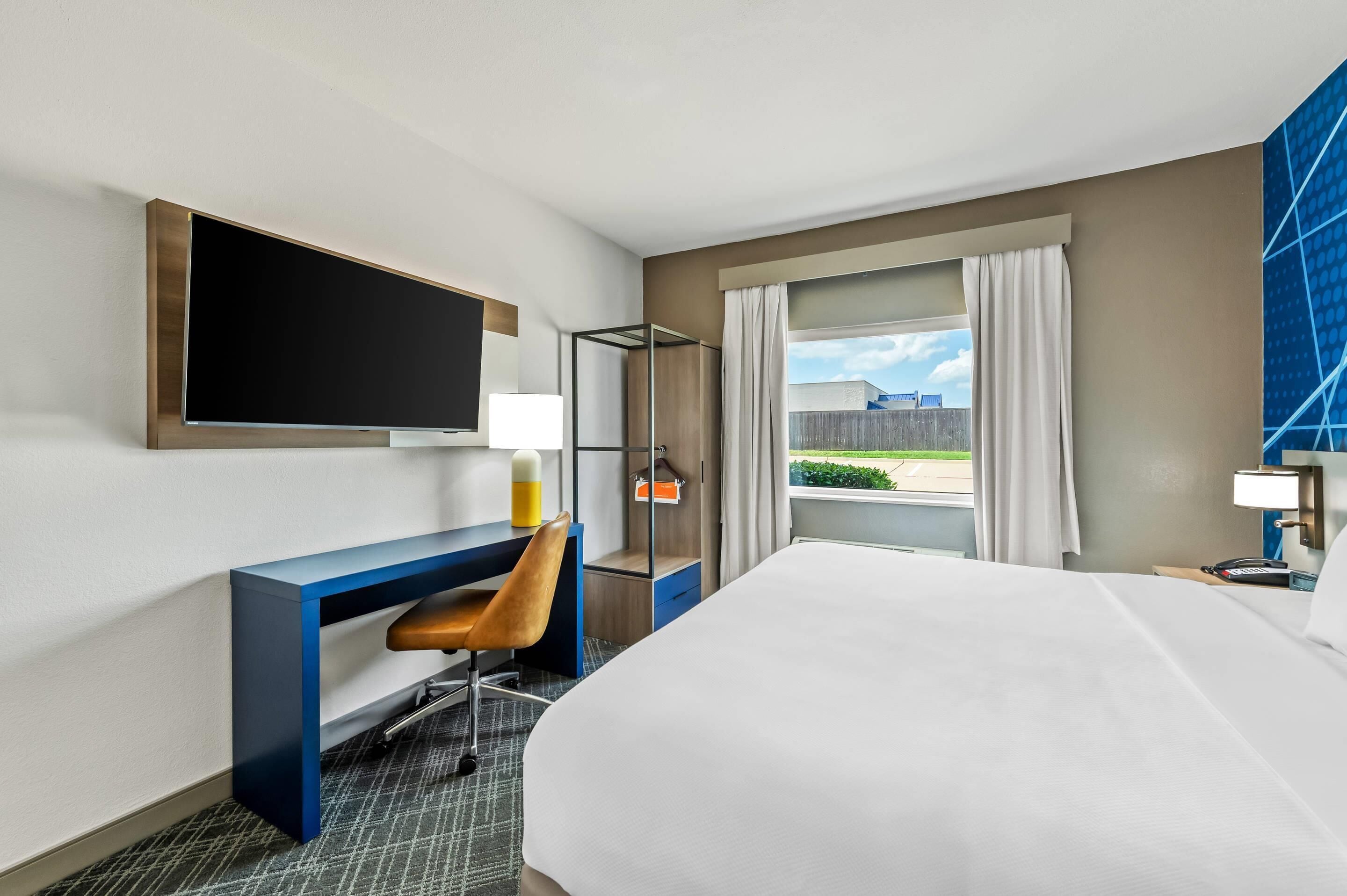 Habitación de hotel con cama, escritorio, silla, televisión y ventana con cortinas.