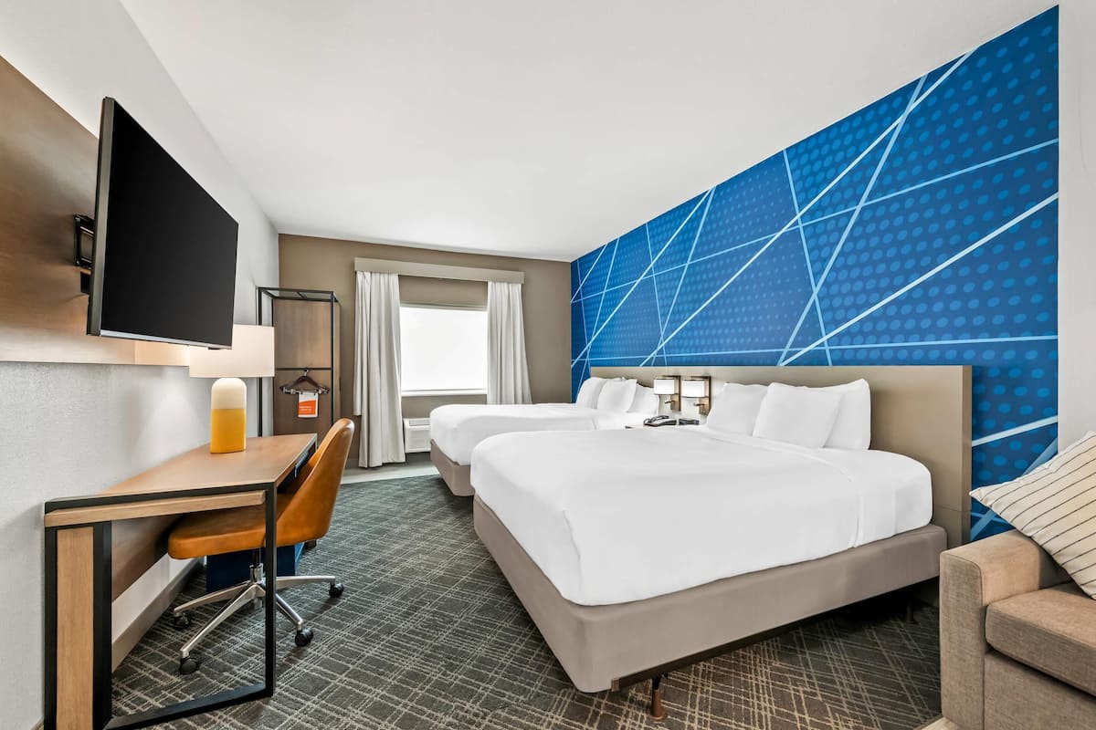 Quarto de hotel com duas camas, uma escrivaninha com cadeira, televisão de tela plana, uma janela grande com cortinas e um design geométrico azul na parede.