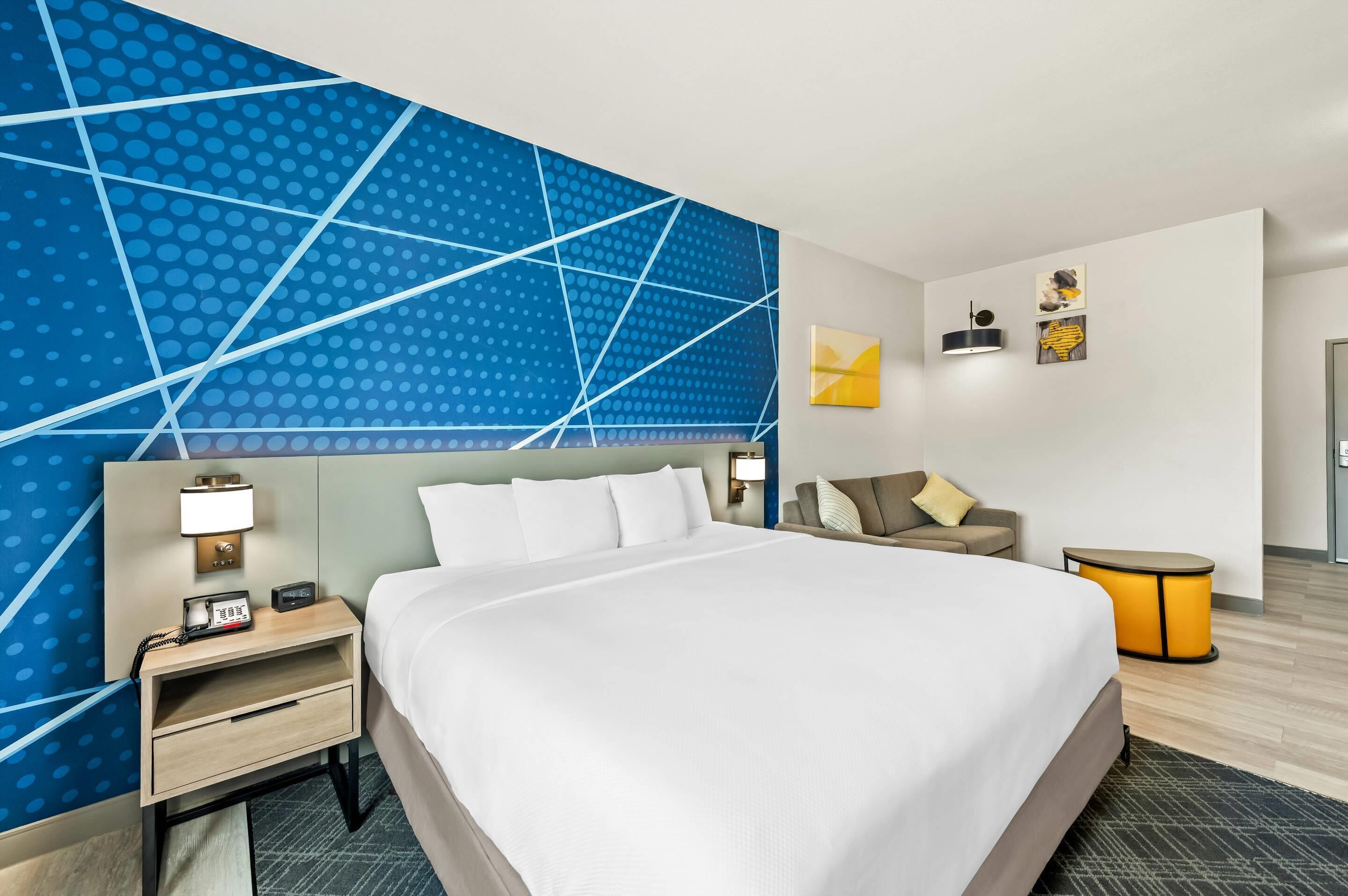 Una habitación de hotel con una cama grande, una mesita de noche con lámpara, una pequeña mesa y una pared con diseños geométricos.