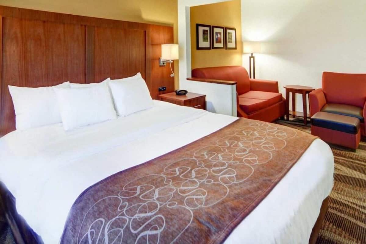 Quarto de hotel com uma cama grande, cabeceira de madeira, uma escrivaninha, uma cadeira e uma mesinha.