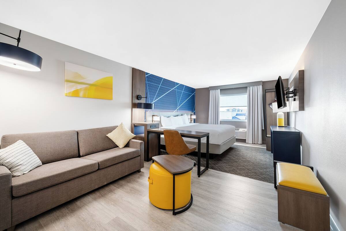 Quarto de hotel moderno com sofá, um banquinho redondo amarelo, mesa de madeira, cama, escrivaninha e televisão fixada na parede.