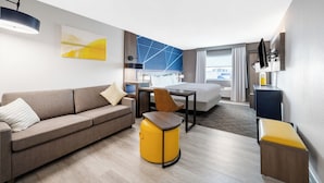 Moderni hotellihuone, jossa on sohva, pieni pyöreä keltainen rahi, puinen pöytä, sänky, työpöytä ja seinälle asennettu televisio.