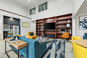 Una sala de estar moderna con sofá, mesa de centro y mueble para televisión. Incluye una zona de cocina con refrigerador y máquina expendedora.