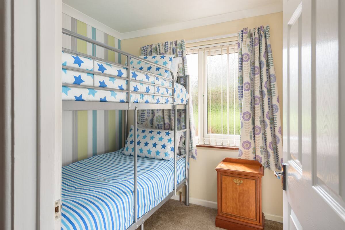 A bunk bed room with a window and curtains, a wooden nightstand, and striped walls.