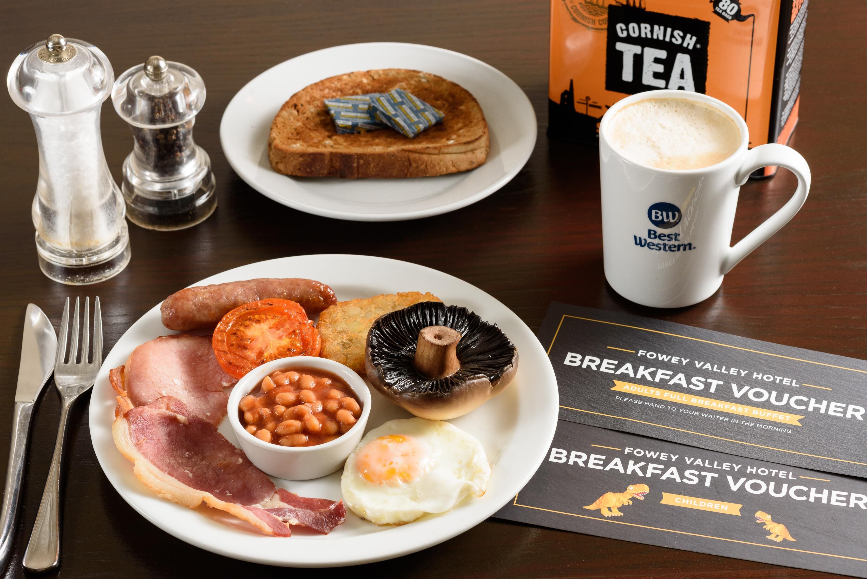 A breakfast plate with eggs, bacon, beans, mushrooms, and toast, accompanied by a mug of coffee and a Cornish Tea box.