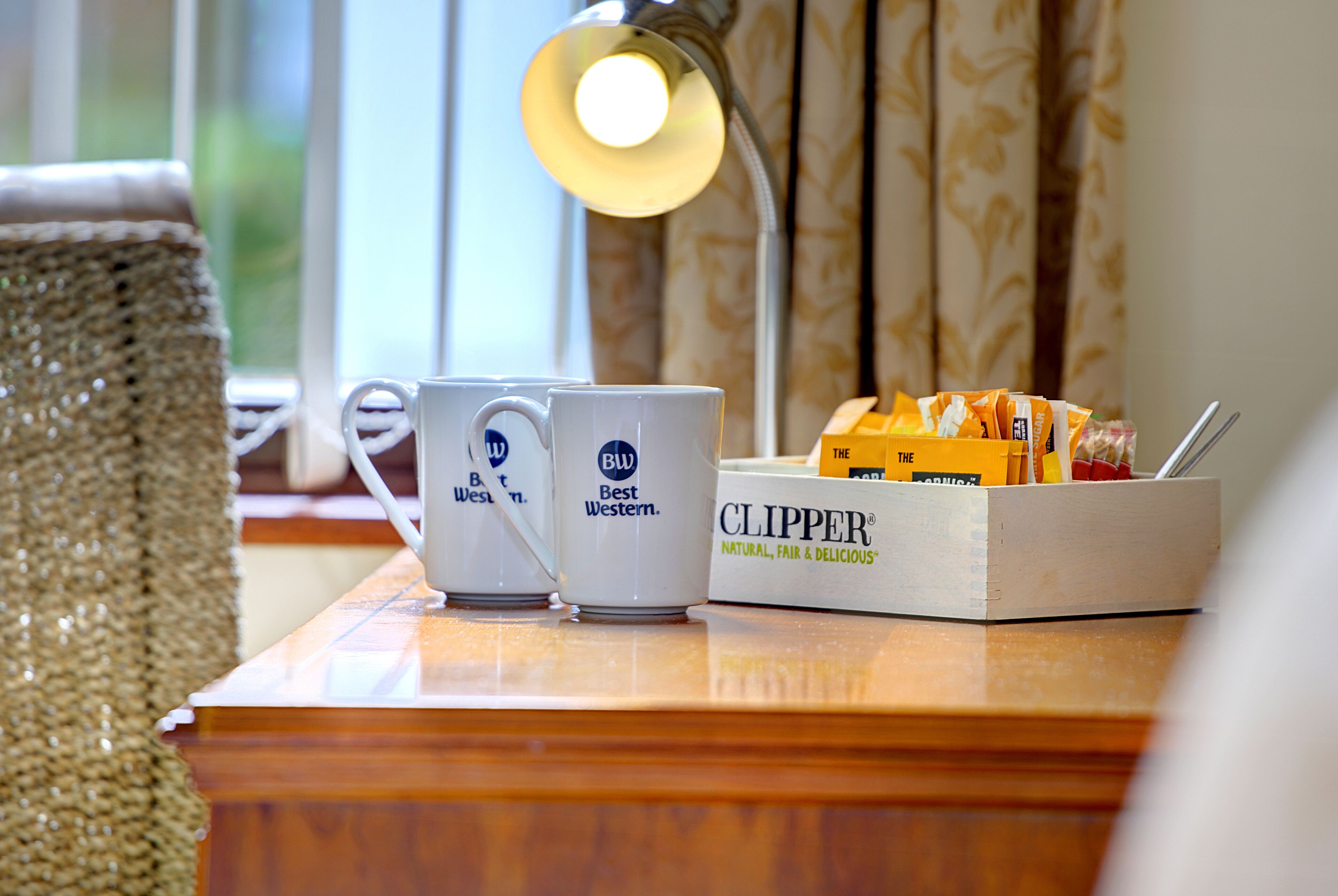 A wooden table with two white mugs labeled "Best Western," a box of Clipper snacks, and a lamp.