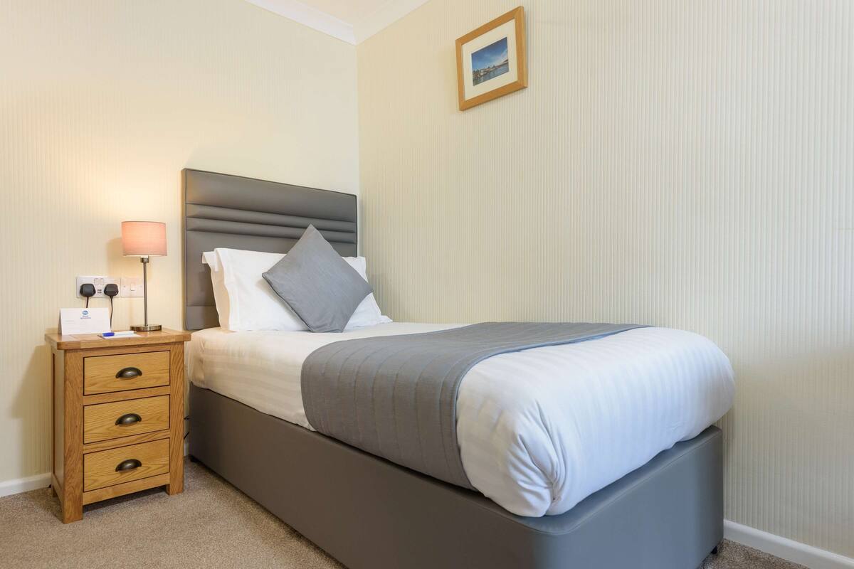 A neatly made bed with a grey headboard, a bedside table with drawers, a lamp, and a framed picture on the wall.