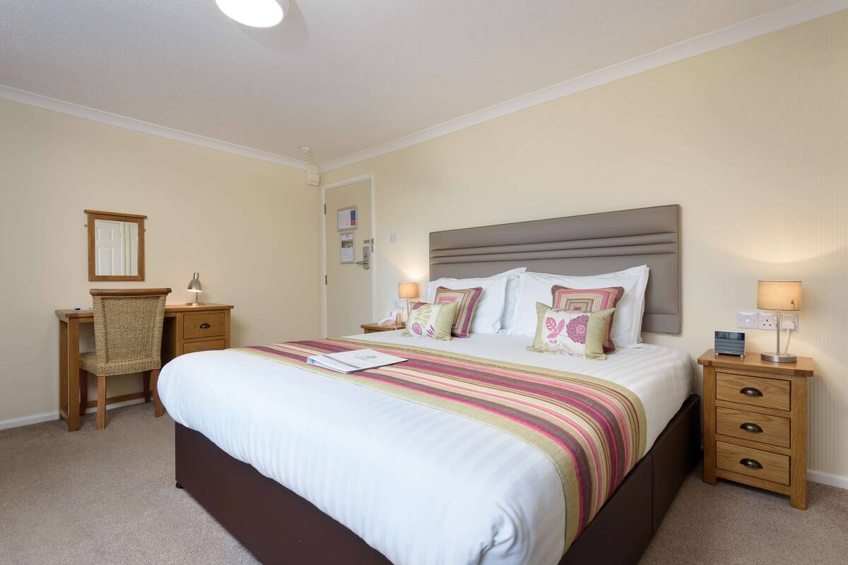 A bedroom with a large bed, a wooden desk, a chair, and a nightstand with a lamp.