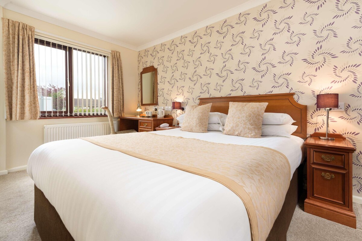 A neatly made bed with a wooden headboard, a bedside table with a lamp, and a window with curtains.