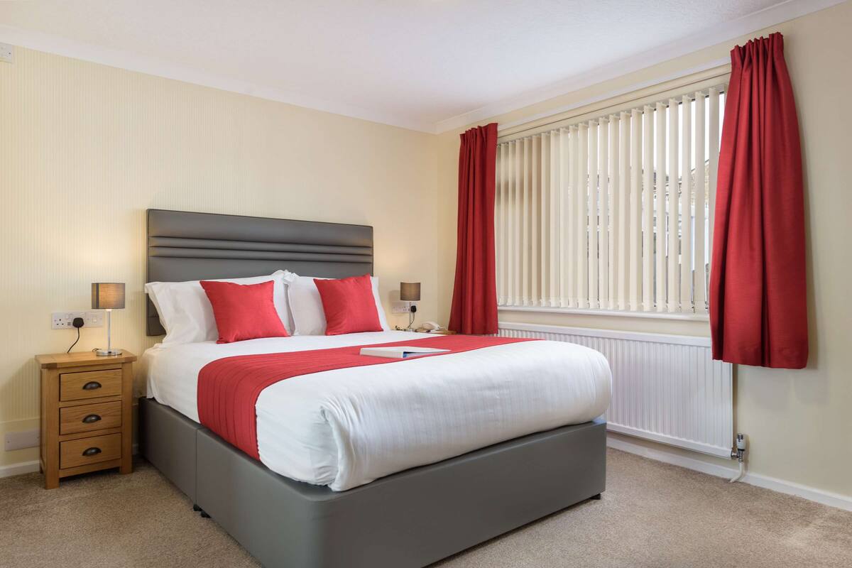 A neatly made bed with a grey headboard, a wooden nightstand with drawers, and a red throw blanket.