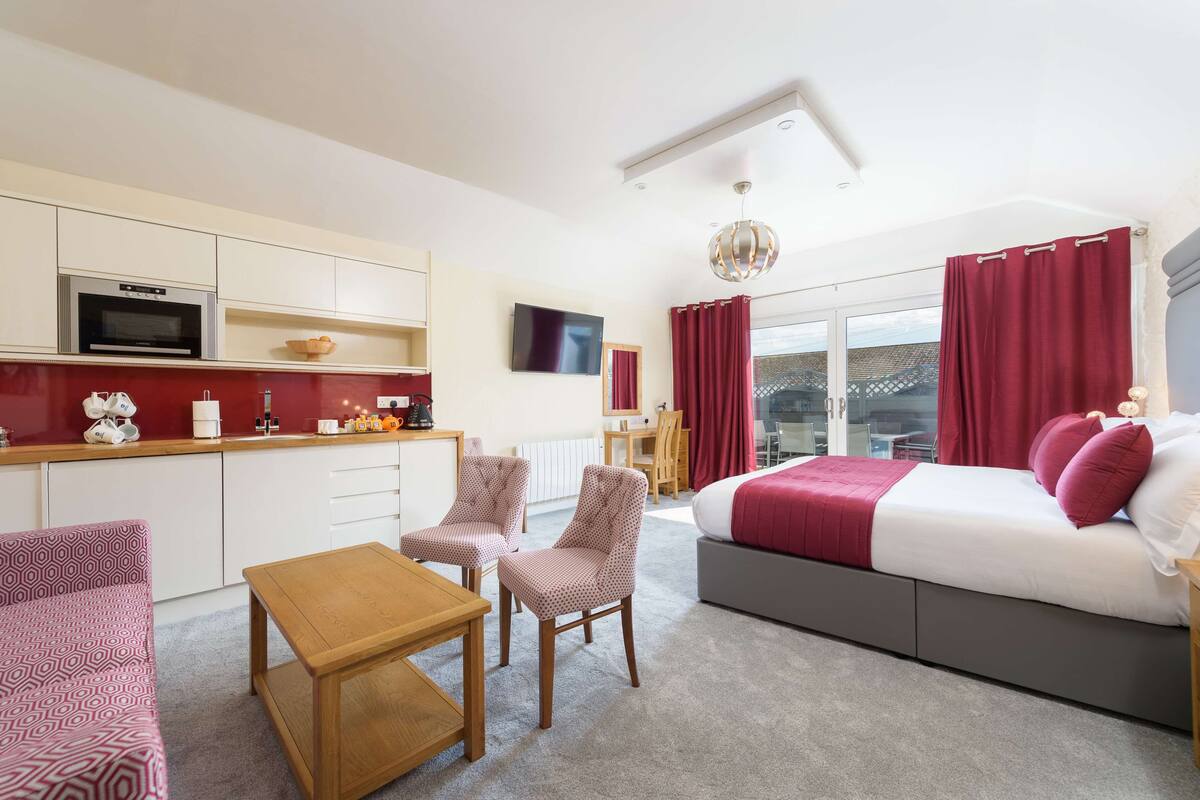 A modern hotel room with a kitchenette, a bed with red bedding, a seating area with two chairs, and a large window with curtains.