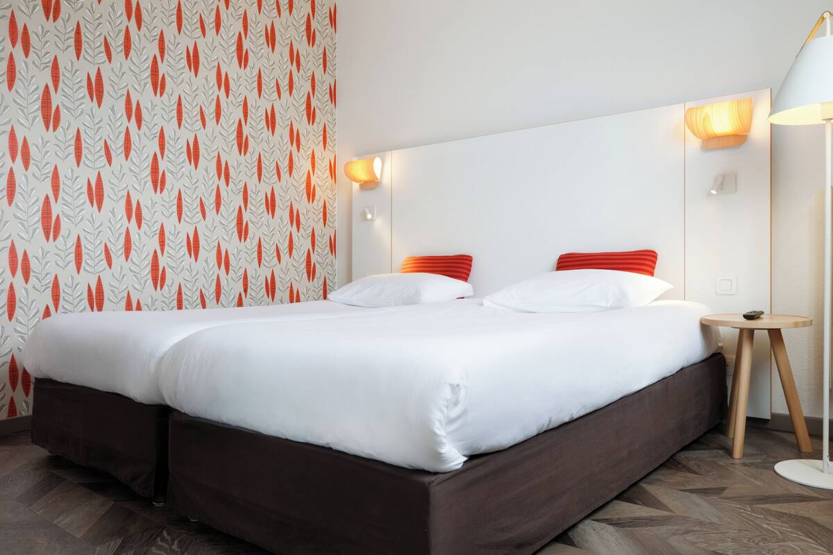 Uma cama de casal com roupa de cama branca e almofadas vermelhas, uma mesa de cabeceira com abajur e um papel de parede estampado com motivos de folhas.