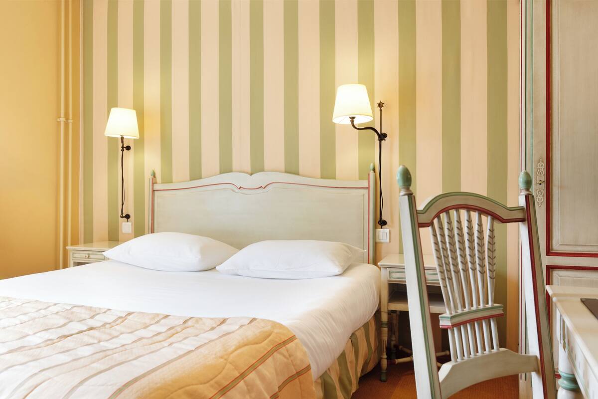 Quarto de hotel com uma cama, duas luminárias de cabeceira, um criado-mudo e uma cadeira.
