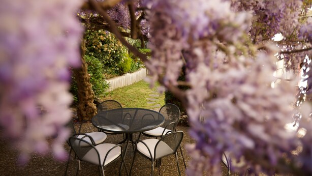 Ein runder Tisch mit Stühlen in einem Gartenbereich im Freien, umgeben von viel Grün und einem weißen Zaun.