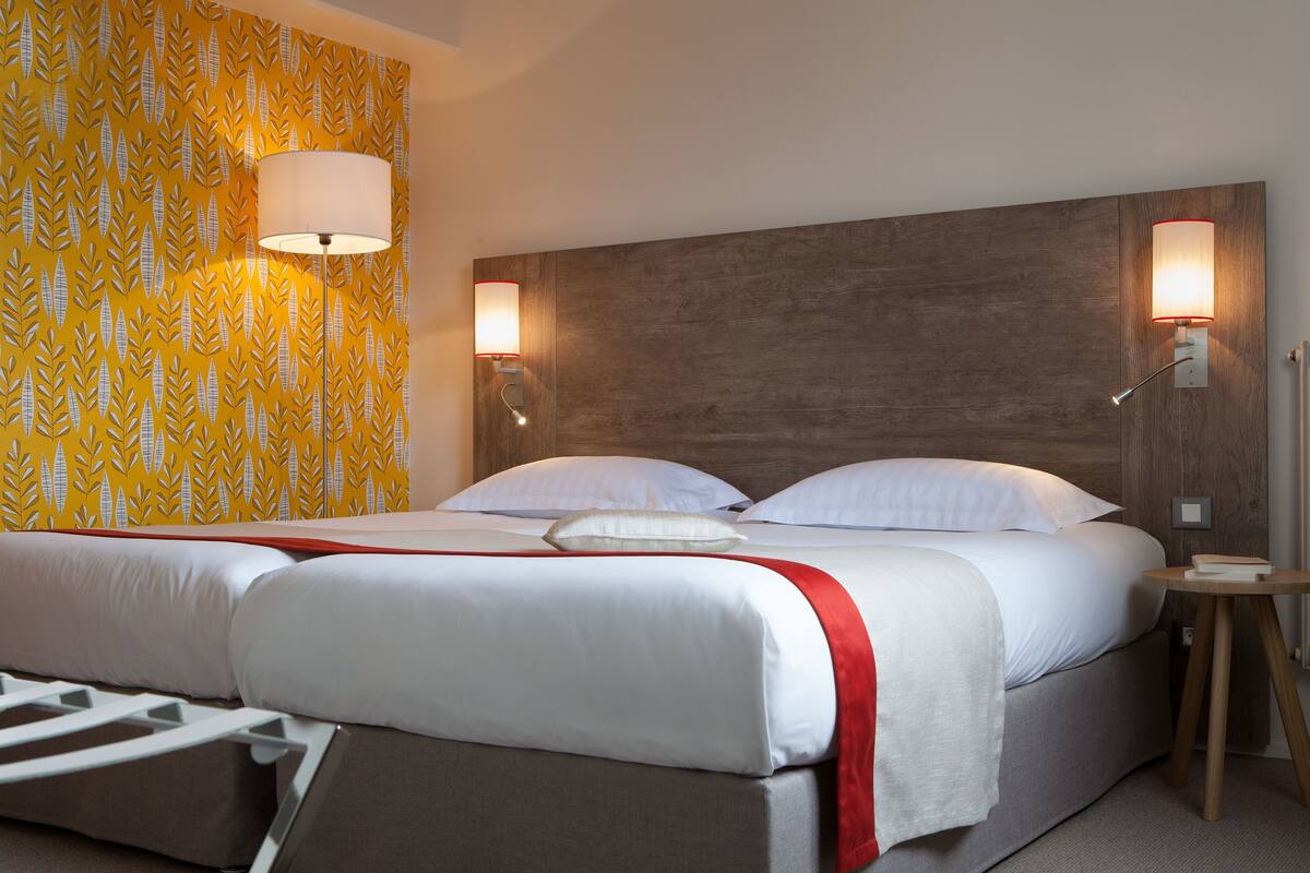 Quarto de hotel com cama, duas luminárias de cabeceira, cabeceira de madeira e papel de parede estampado.