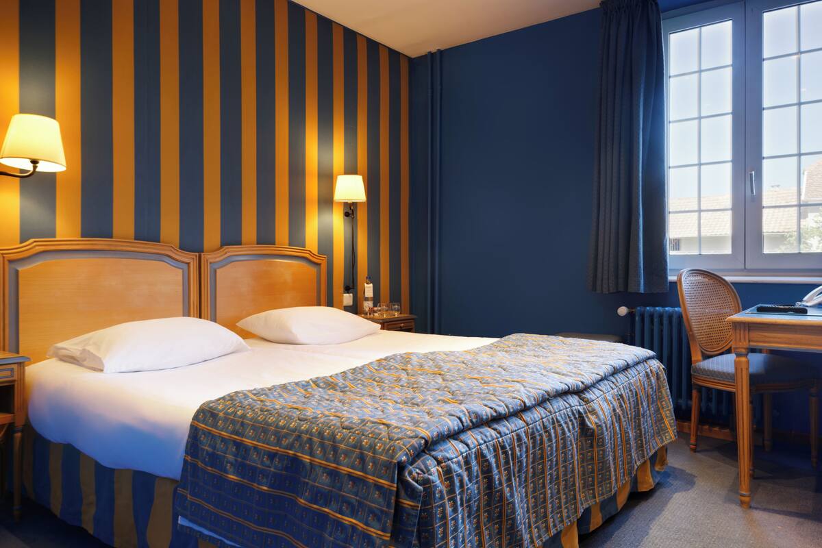 Quarto de hotel com cama, duas luminárias de cabeceira, escrivaninha com cadeira e janela com cortinas.