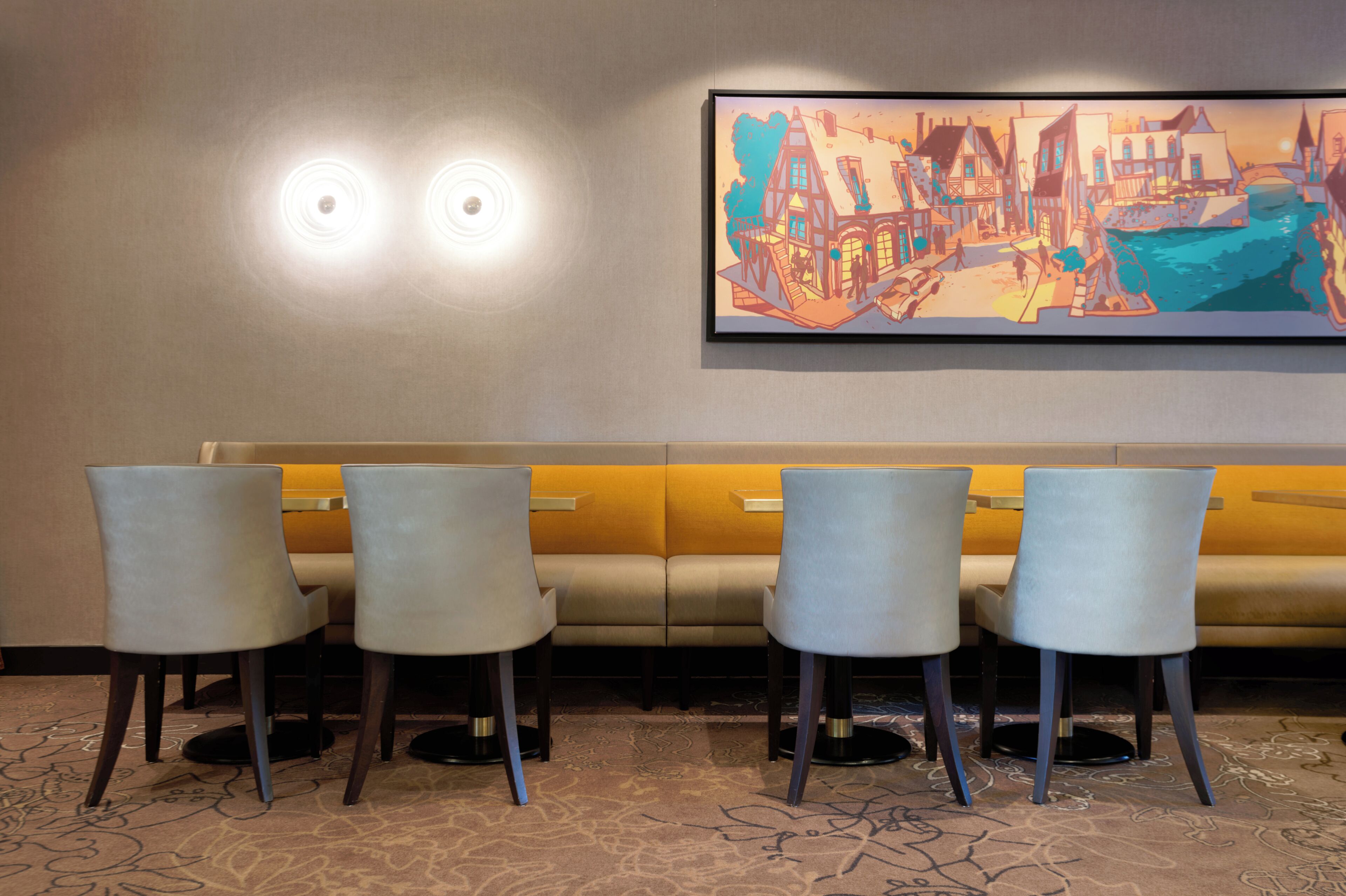 A hotel lobby with a seating area featuring a sofa and four chairs, a large wall-mounted artwork depicting a stylized cityscape, and two ceiling lights.