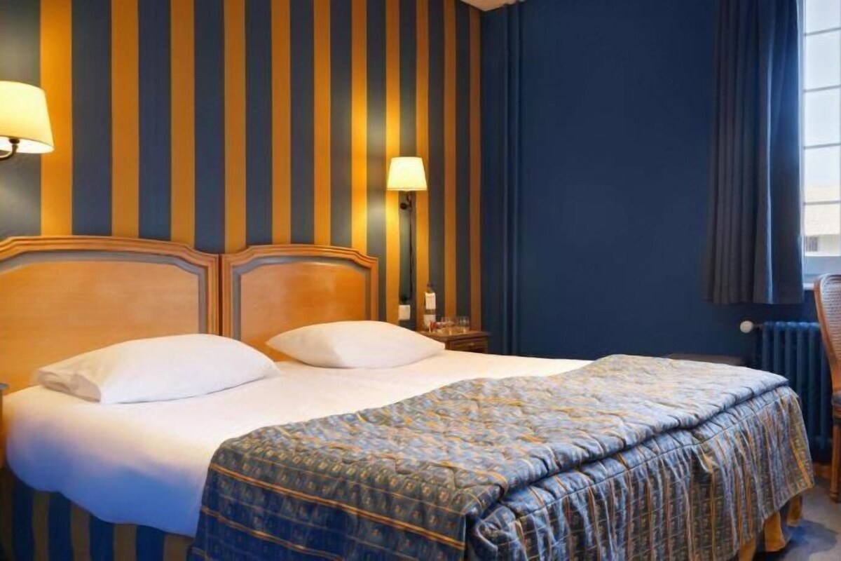 A hotel room with a bed, two bedside lamps, a chair, a table, and a window.