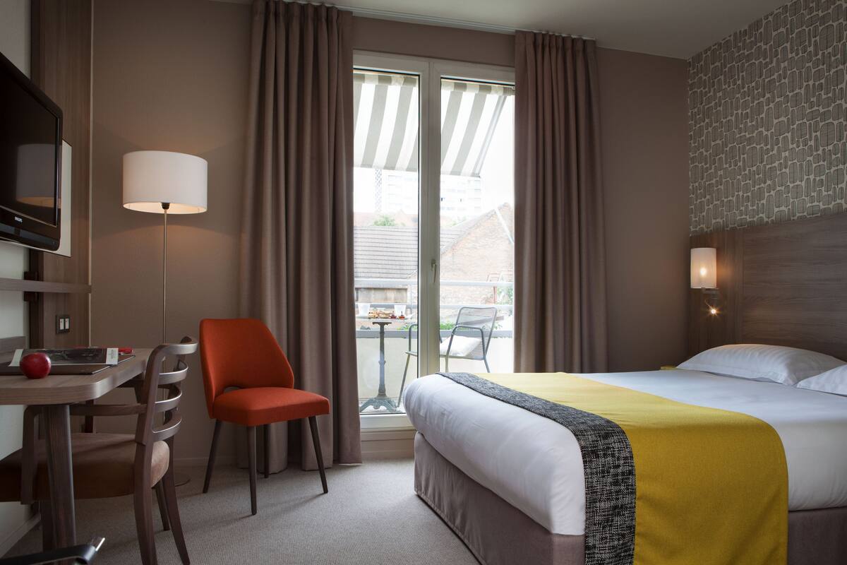 Quarto de hotel com uma cama, escrivaninha, cadeira e janela com cortinas.