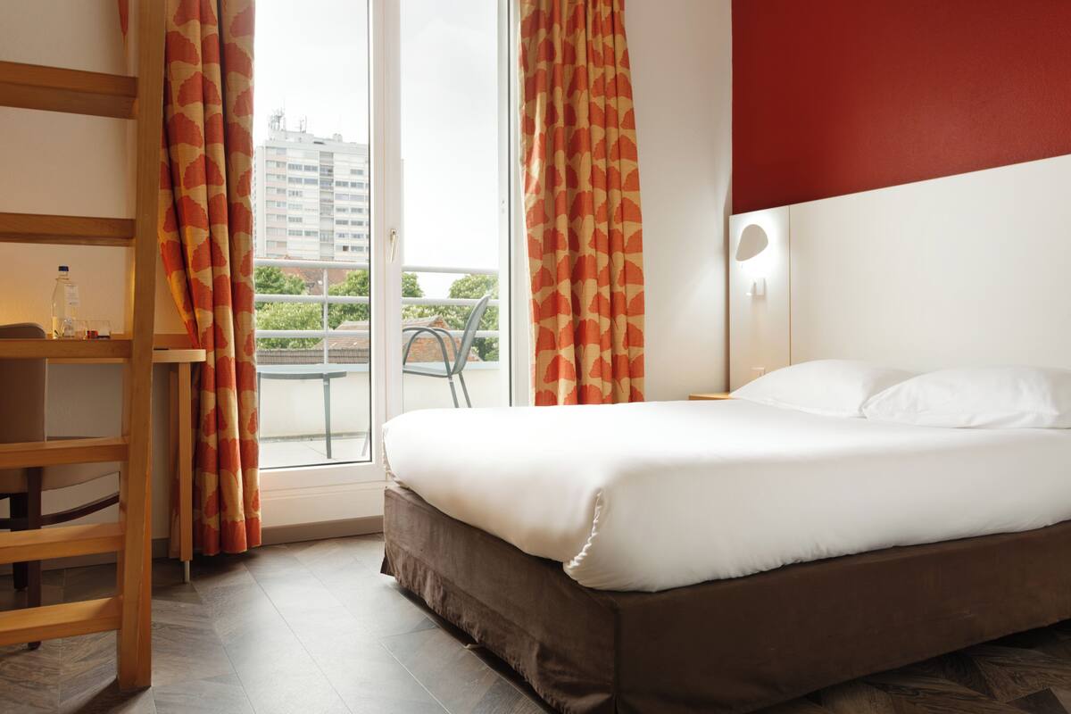 Quarto de hotel com uma cama, uma cadeira de madeira, uma mesa com uma garrafa e cortinas.