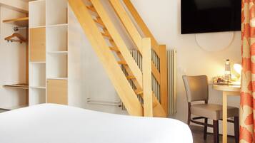 A hotel room with a bed, a wooden staircase, a TV mounted on the wall, a chair, a table with a lamp, and a wardrobe with hanging space.