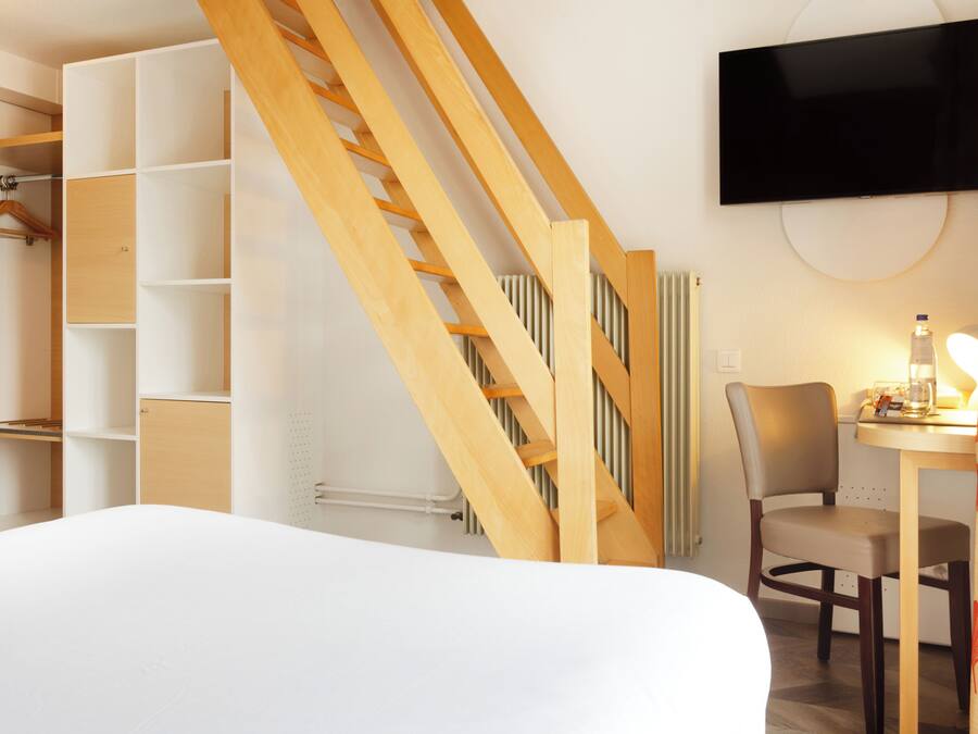 A hotel room with a bed, a wooden staircase, a TV mounted on the wall, a chair, a table with a lamp, and a wardrobe with hanging space.