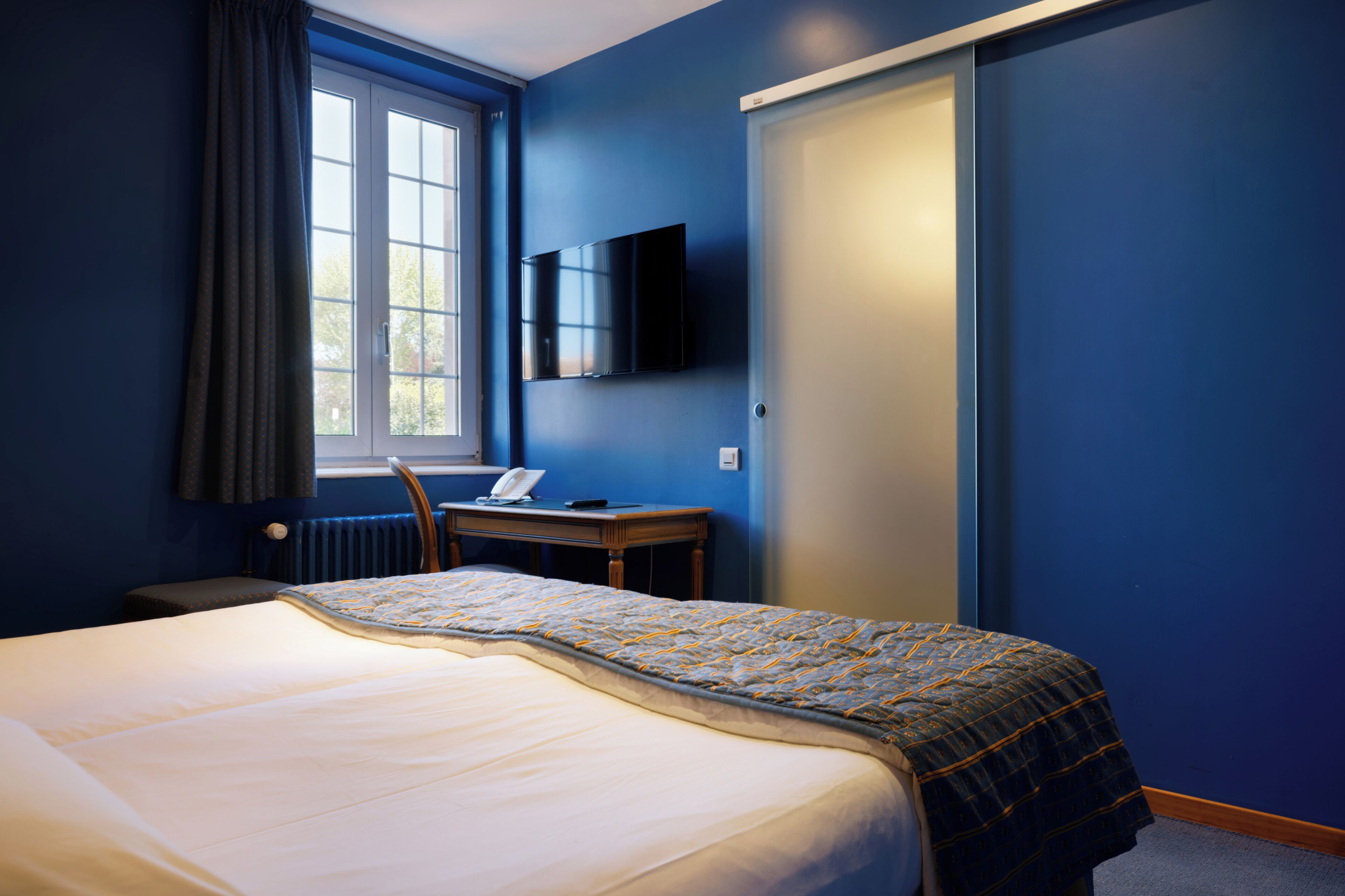 Quarto de hotel com cama, escrivaninha com telefone, televisão na parede e janela com cortinas.