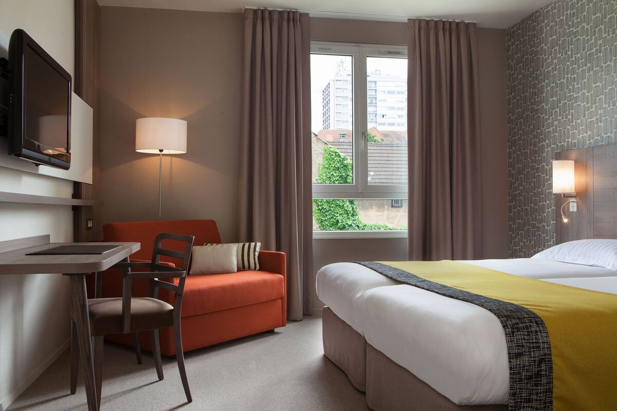 Quarto de hotel com cama, escrivaninha, cadeira, sofá e janela com cortinas.
