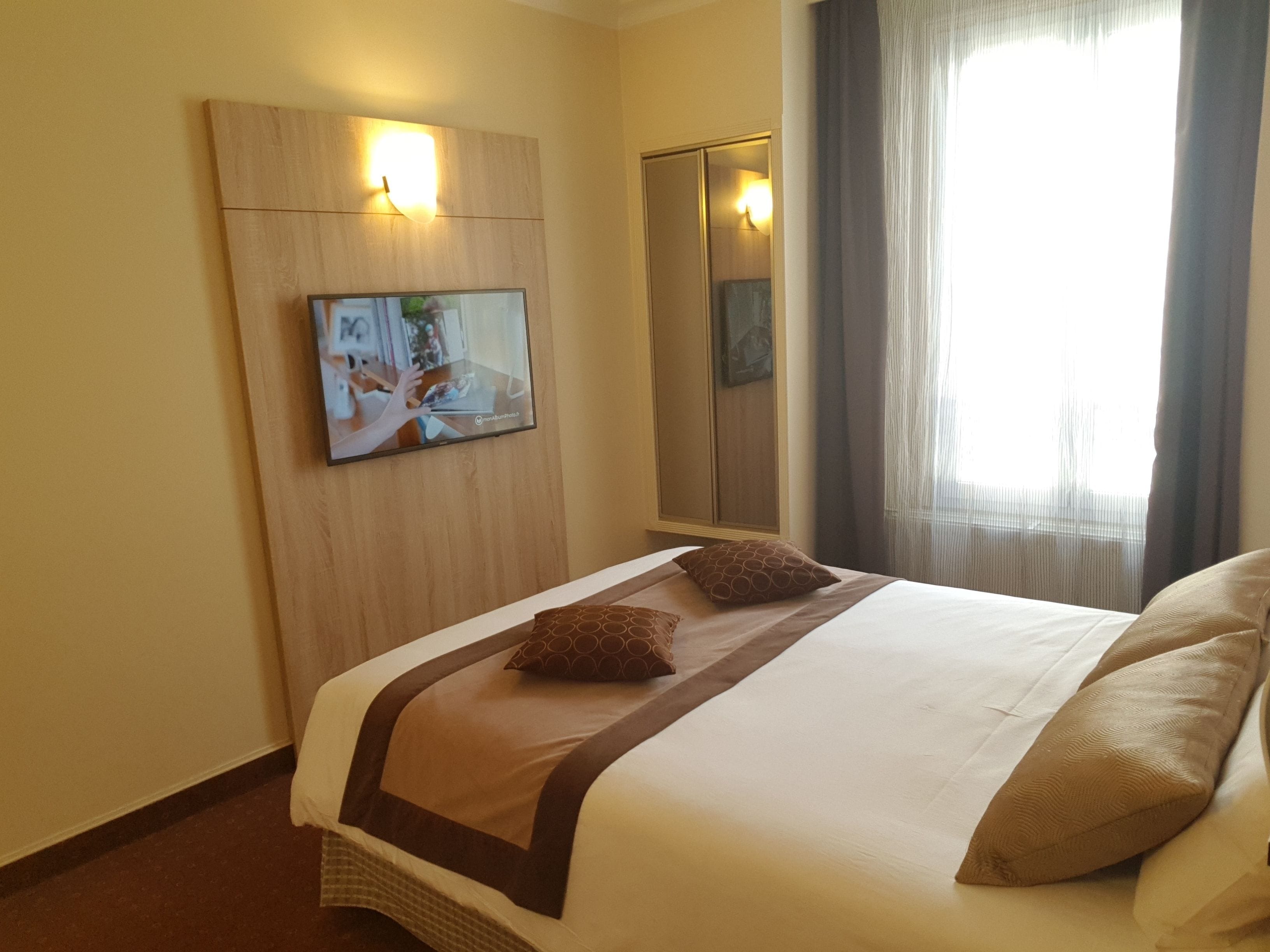 A hotel room with a bed, a television mounted on the wall, a wardrobe, and a window with curtains.