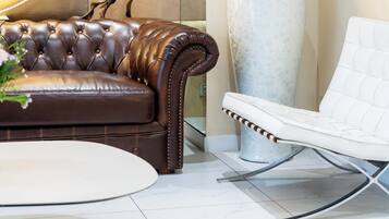 A modern living room with a brown leather sofa, a white armchair, a large vase with dried plants, and a patterned wall.