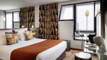 A modern hotel room with a large bed, a bedside table with a lamp, a mirror, and a window with curtains.