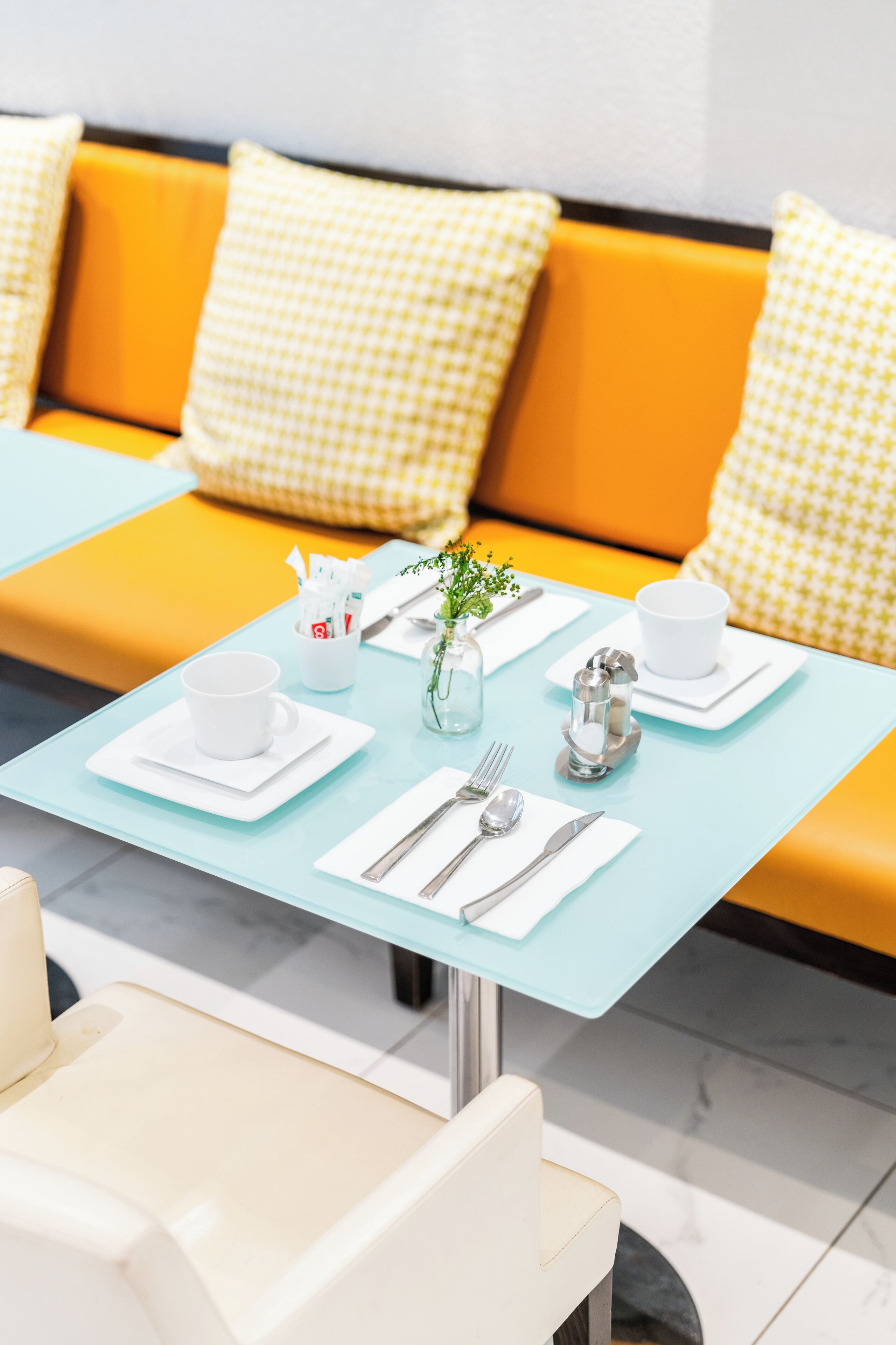 Un comedor con una mesa azul claro, platos blancos, cubiertos y una taza blanca. Hay un pequeño jarrón con una planta sobre la mesa.