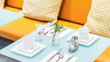 Un comedor con una mesa azul claro, platos blancos, cubiertos y una taza blanca. Hay un pequeño jarrón con una planta sobre la mesa.