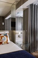 A modern hotel room with a large bed, a glass side table, a mirror, and a view of the outdoors through a window with curtains.