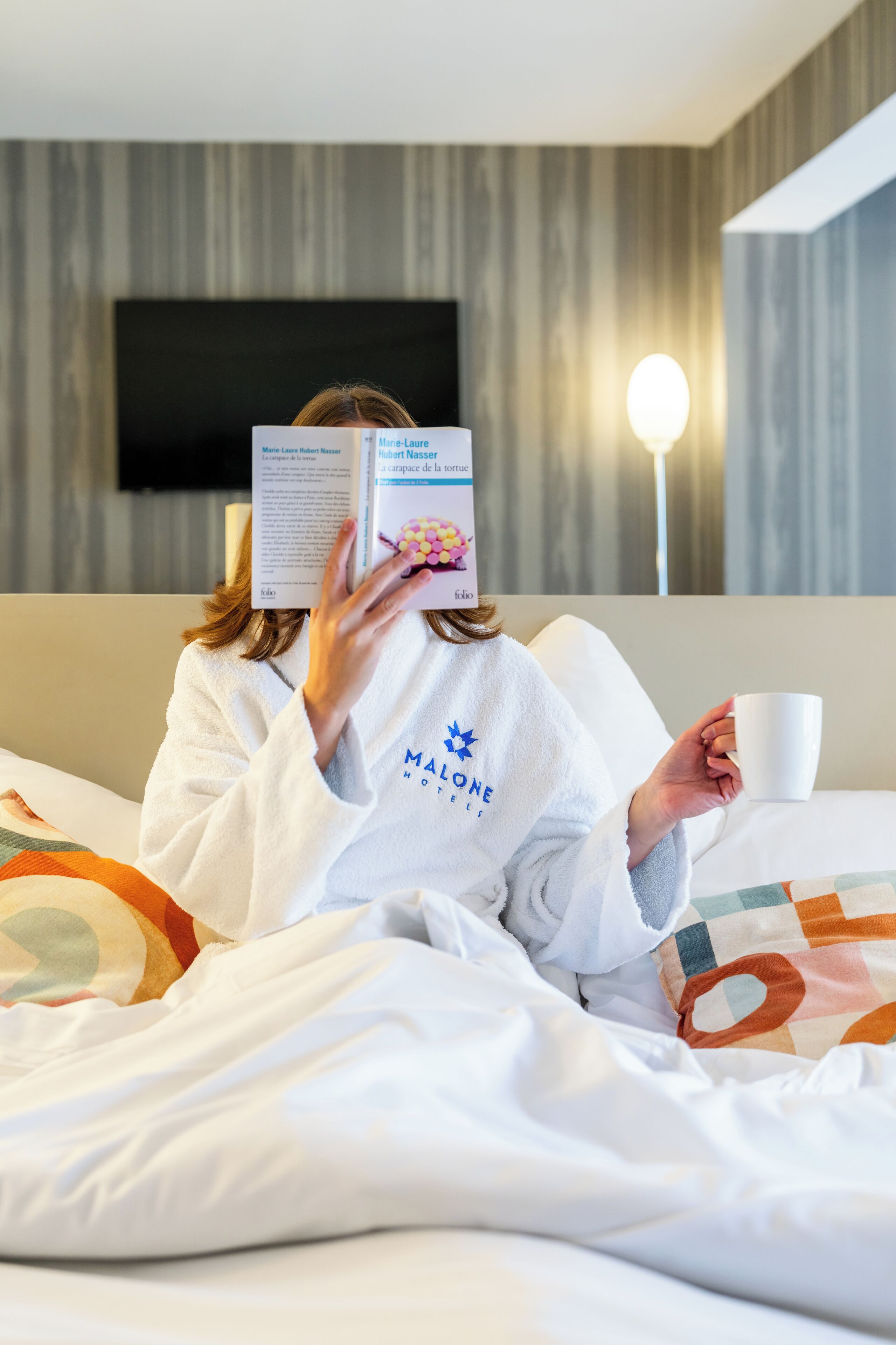Une personne est assise dans une chambre d’hôtel, un livre à la main, devant un lit, des oreillers et un téléviseur fixé au mur.