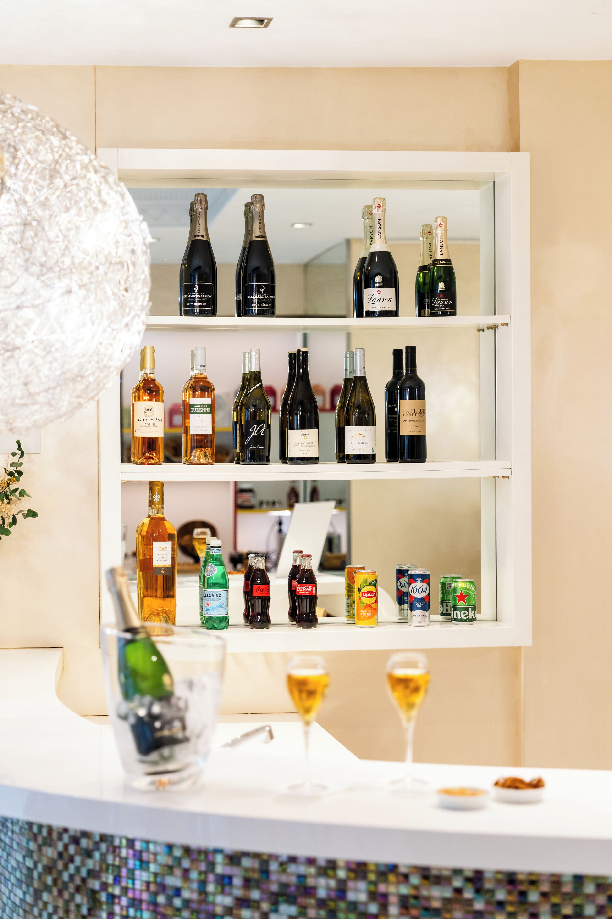 Un coin bar avec des étagères remplies de bouteilles de différentes sortes d’alcool, un verre décoratif et deux verres contenant une boisson dorée.